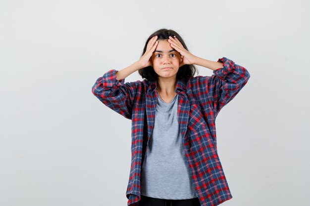 Cute teen girl in checked shirt holding hands on head and looking confused , front view.