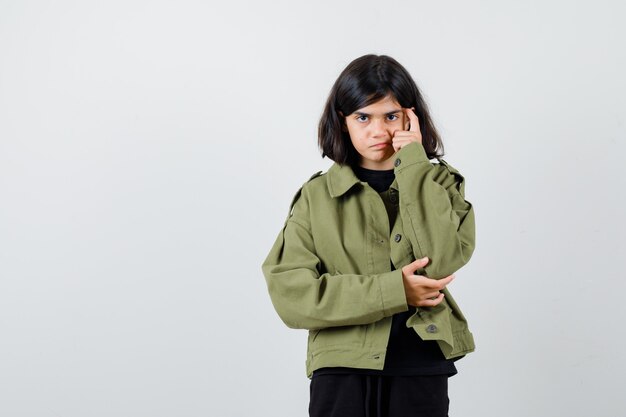 Cute teen girl keeping finger on temples in army green jacket and looking intelligent , front view.