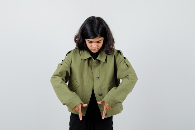 Симпатичная девочка-подросток, указывая вниз в армейской зеленой куртке и выглядящая сосредоточенной. передний план.