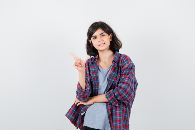 Cute teen girl pointing at upper left corner in checked shirt and looking confident , front view.