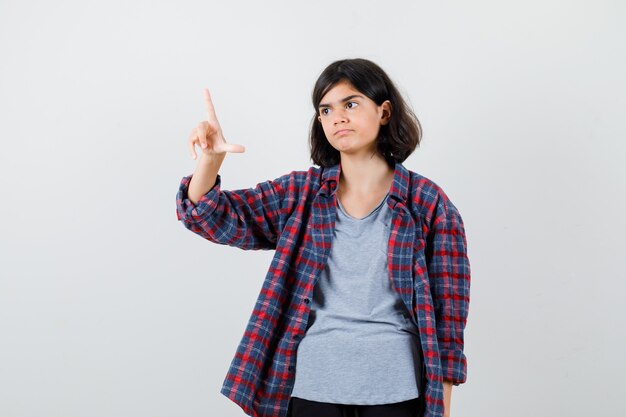 Cute teen girl showing loser sign while looking away in checked shirt and looking cheerless , front view.
