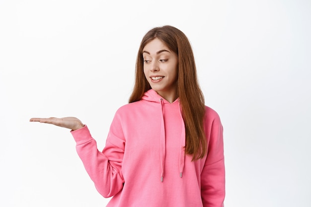 Cute teen girl student, holding copyspace on palm, display product in her hand against white wall, smiling and looking at object, white wall