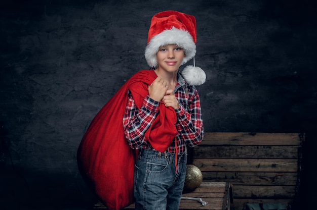 산타 모자를 쓴 귀여운 10대 소년이 새해 선물 자루를 들고 있습니다.