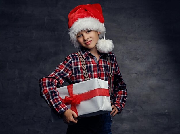 Cute teenage boy in Santas' hat dressed in a plaid shirt holds big present gift box.