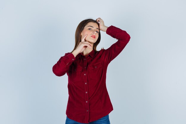 Cute teenage girl keeping hand on head, holding finger on cheek in burgundy shirt and looking delicate , front view.