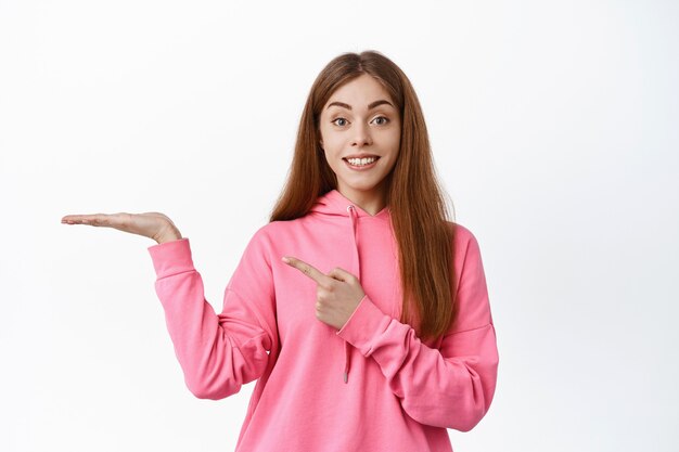 Cute teenage girl pointing finger at empty hand, display copyspace logo on her palm, smiling at front, standing over white wall