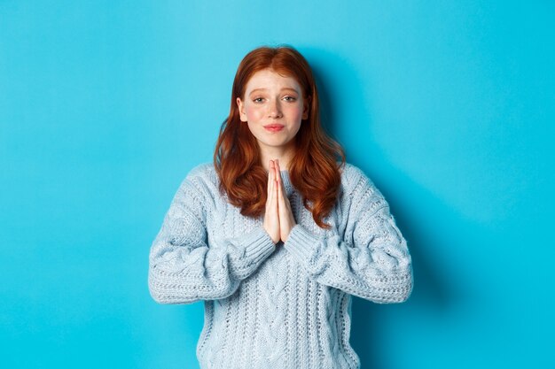Cute teenage redhead girl asking for help, smiling while begging for favour, need something, standing over blue background