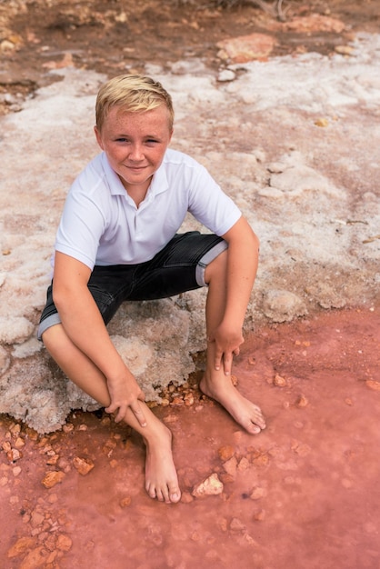 Cute teenager blonde boy wearing summer clothes sitting on shore of an amazing pink lake