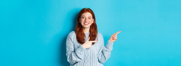 Free photo cute teenager girl with red hair and freckles pointing fingers left at logo and smiling showing adve