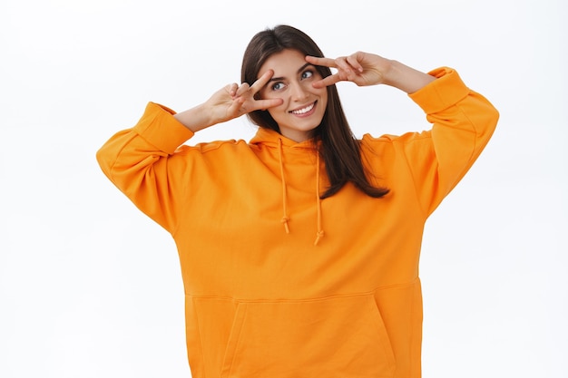 Cute tender feminine caucasian woman in orange hoodie, show peace signs over eyes, tilt head and looking left happy, smiling broadly express positive vibe, cheer and joy, white wall