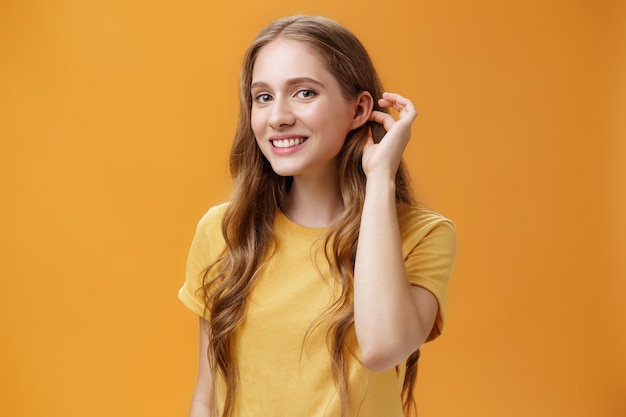 Cute and tender flirty young woman with cute wavy hairstyle flicking hair strand behind ear gazing at camera with timid sensual smile glancing with admiration at guy over orange background. Copy space