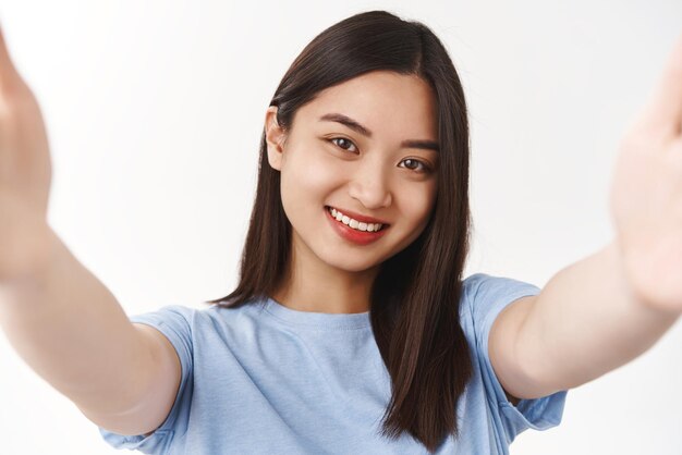 Cute tender smiling sensual asian brunette hold camera hands stretched forward taking selfie digital tablet talking videocall grinning toothy express positive good mood white background