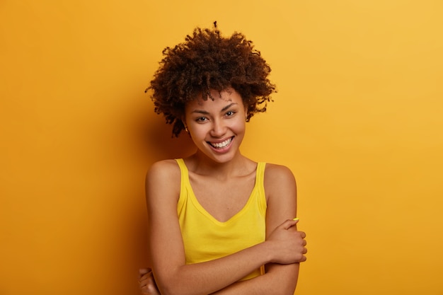 Cute tender young woman keeps hands crossed over body, smiles sensually and gazes , has natural curly hair, enjoys awesome moment in life, poses against yellow wall, has pleasant talk