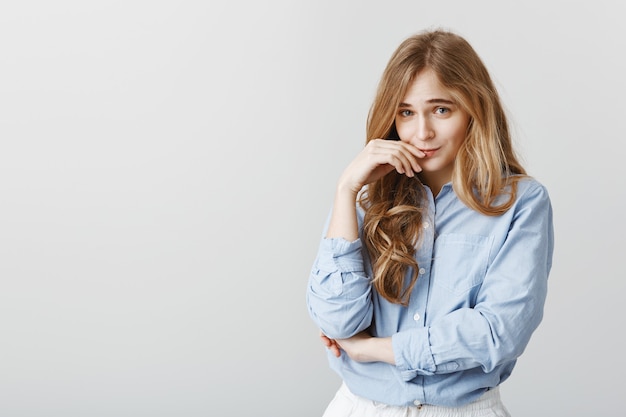 Cute timid female student in blue-collar shirt, holding hand near lip and lifting eyebrows, making gentle expression, pretending innocent