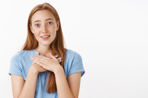 Cute touched ginger girl with freckles holding palms on heart melting from charming surprise smiling and gazing with admiration and affection being grateful over gray wall