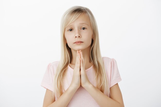 Cute upset girl begging and apologizing. Portrait of serious moody blond daughter in adorable outfit, holding hands in pray with clenched palms, hoping for forgiveness over gray wall