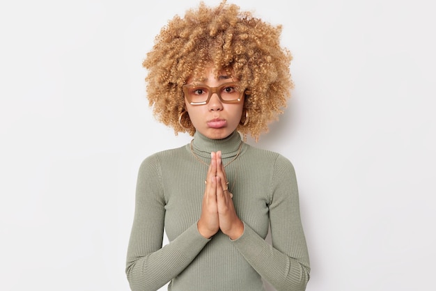 Cute upset young woman with curly hair keeps palms pressed together begs and shows beg gesture looks hopeful wears spectacles casual turtleneck isolated over white background. Please help me