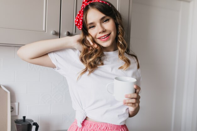 Милая белая девушка в повседневной футболке, стоя на кухне с чашкой чая. Крытый выстрел расслабленной женской модели, пьющей кофе утром.