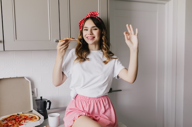 Cute white girl with ribbon in hair sitting in kitchen with smile. Gorgeous european lady eating pizza with happy face expression.