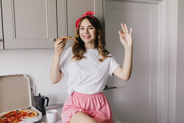 Cute white girl with ribbon in hair sitting in kitchen with smile. Gorgeous european lady eating pizza with happy face expression.