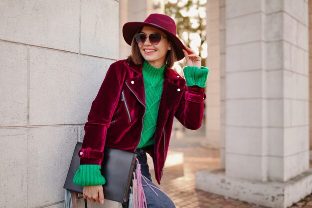 Cute woman in autumn style trendy outfit walking in street wearing purple velvet jacket, sunglasses and hat