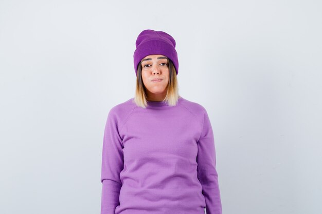 Cute woman blowing cheeks in sweater, beanie and looking puzzled , front view.
