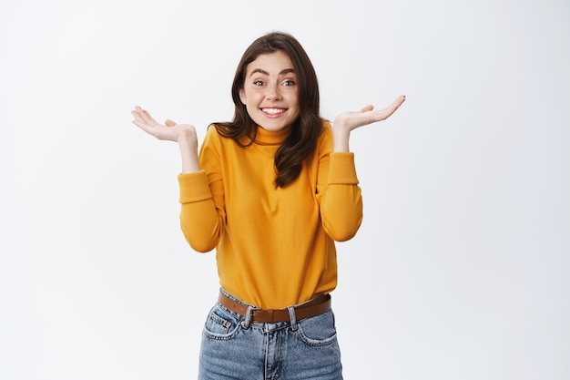 Cute woman dont know anything, raising hands up and shrugging with silly smile, have no idea, say sorry for being unaware, standing against white wall