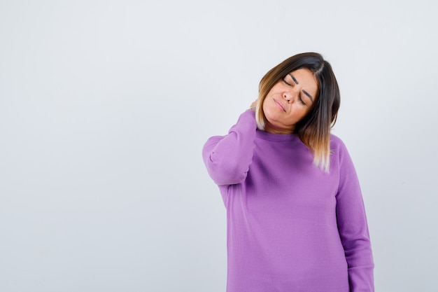 Милая женщина чувствует боль в шее в фиолетовом свитере и выглядит усталой, вид спереди.
