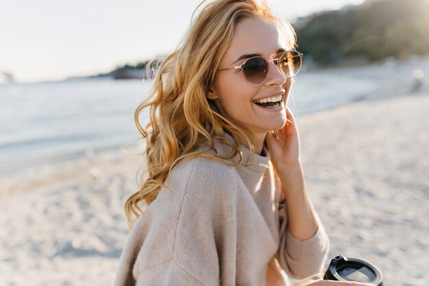 Cute woman genuinely laughs, relaxing in beach. Blinde woman in glasses and sweater holds cup of coffee.