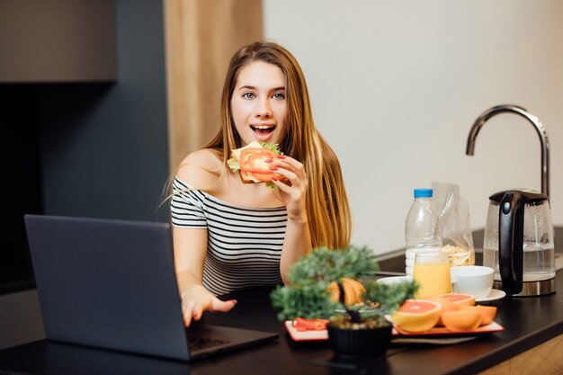 맛있는 샌드위치를 먹고 부엌에서 전화를 사용하면서 건강한 아침 식사를 하는 귀여운 여자