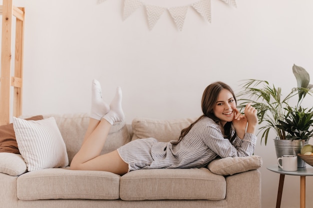 Cute woman laughs and looks into front while lying on sofa in cozy living room