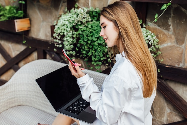 Милая женщина делает покупки в интернете с помощью серого ноутбука с телефоном на роскошной террасе. покупка онина.