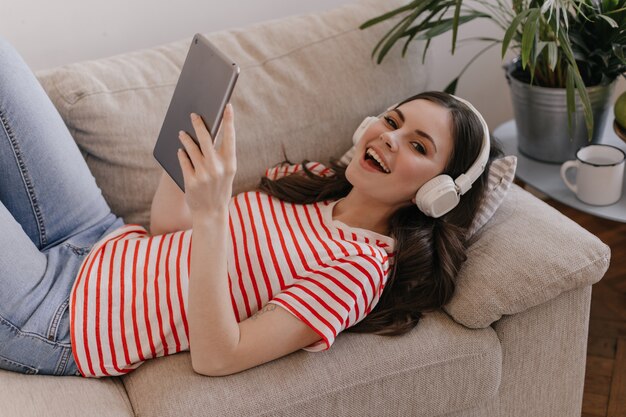 Cute woman in new white headphones is resting on soft sofa and having fun