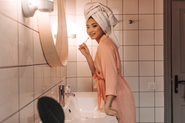 분홍색 가운에 귀여운 여자가 치아를 청소하고 욕실 거울 벽에 정면으로 보입니다.