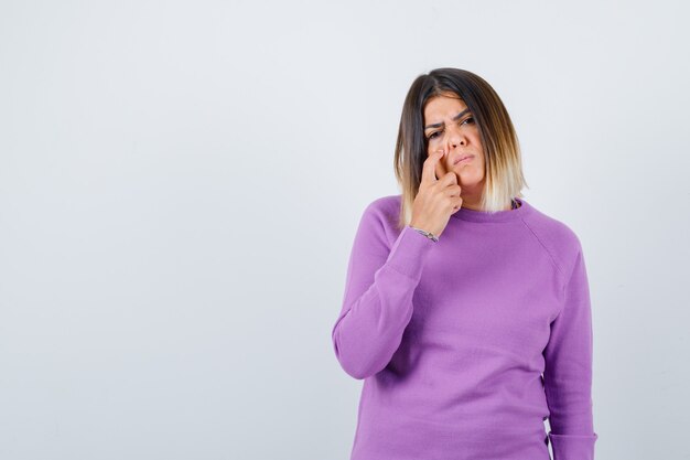 Cute woman in purple sweater holding finger on cheek and looking gloomy , front view.