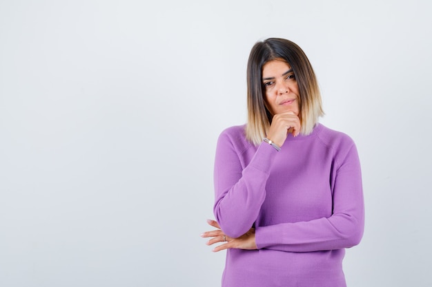 Foto gratuita donna carina in maglione viola con la mano sul mento e dall'aspetto pensieroso, vista frontale.