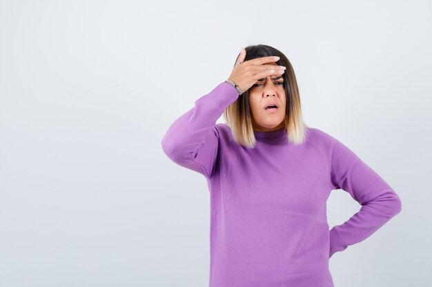 보라색 스웨터를 입은 귀여운 여성이 이마에 손을 대고 우울한 표정을 짓고 있습니다.