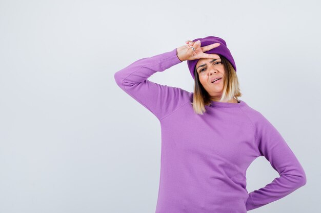 Cute woman showing victory gesture in sweater, beanie and looking proud , front view.