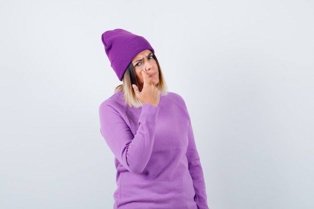 Free photo cute woman in sweater, beanie pointing at her eyelid and looking upset , front view.