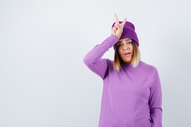 Cute woman in sweater, beanie showing peace gesture and looking wondered , front view.