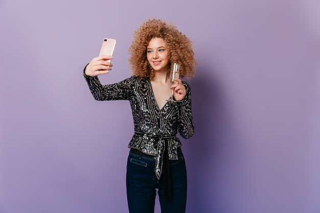 Free photo cute woman with blond curls dressed in black sequined top holding glass of champagne and making selfie on purple space.