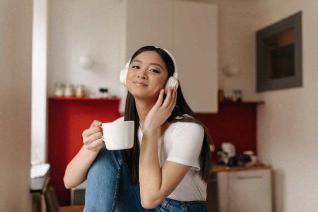 Cute woman with smile looks into front, listens to music on headphones and holds white cup on background of kitchen
