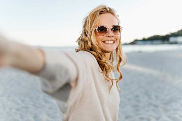 Cute woman with wavy blind hair sincerely smiles and takes selfie at sea.