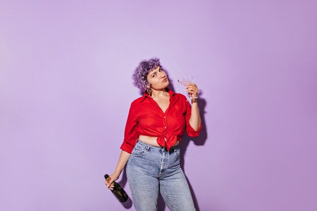 Cute wonderful woman in red stylish shirt holds glass and bottle of white wine and on lilac.