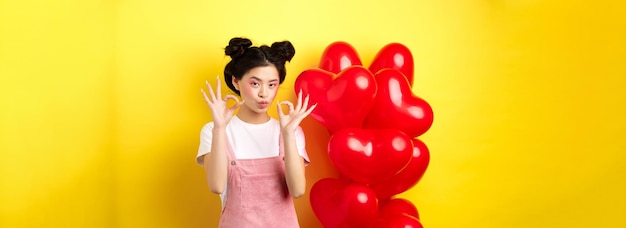Foto gratuita carina giovane donna asiatica in abiti eleganti e trucco che mostra segni ok vicino al cuore di san valentino