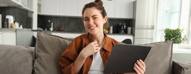 Free photo cute young brunette woman holding digital tablet in hand sitting on couch smiling and laughing at