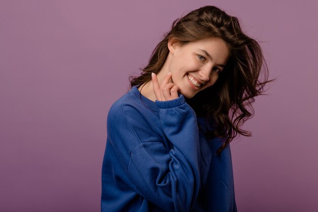 Cute young caucasian brunette girl in blue hoodie looks at camera on purple background Lifestyle concept