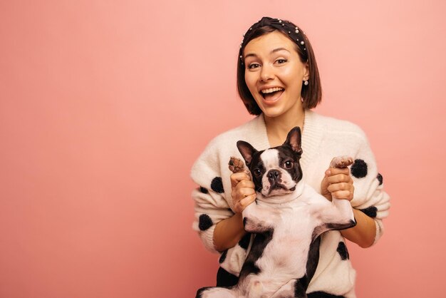 Cute young caucasian brunette girl holding on to paws of dog in front camera on pink background Good relationship between animals and people concept
