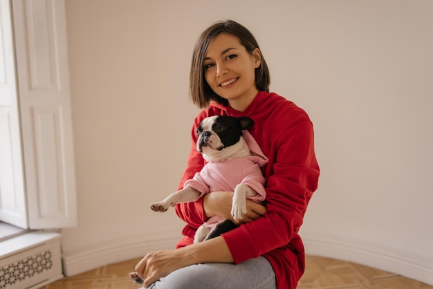 Cute giovane ragazza bruna caucasica tiene il bulldog francese sulle braccia in una stanza spaziosa amore per gli animali domestici gioia e tenerezza