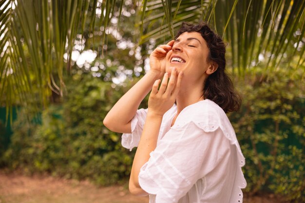 Cute young caucasian brunette girl in white summer clothes smiles with her eyes closed touching face outdoors